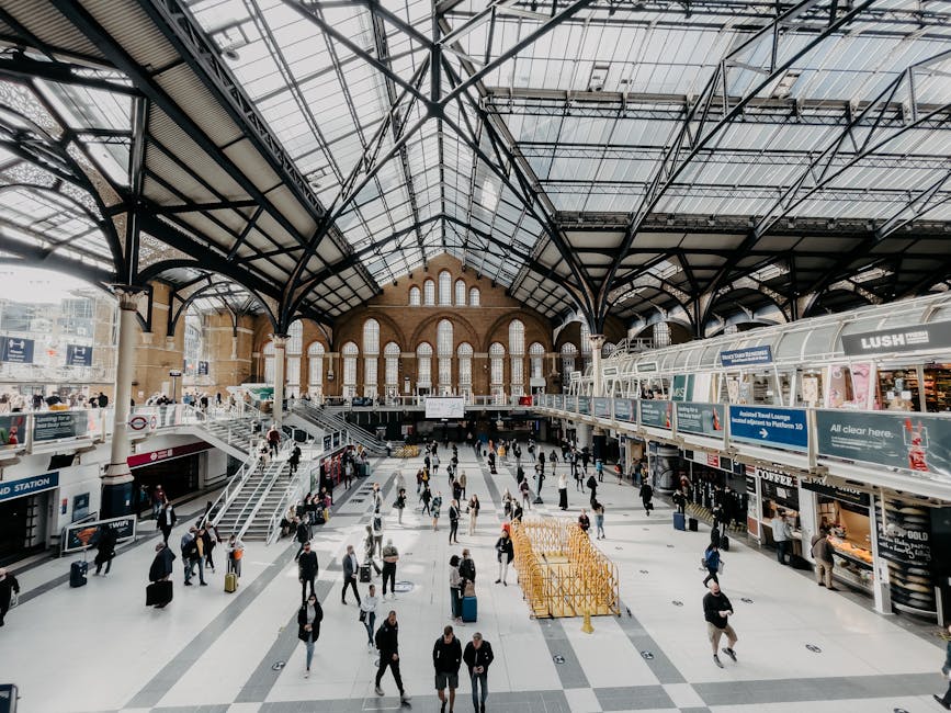 The busy interior of Fulham Broadway station, featuring a high glass and metal vaulted ceiling with intricate structural framework, large arched windows allowing natural light to illuminate the spacious concourse. The polished tiled floor appears clean and reflective, with groups of travelers walking, standing, and waiting, some pulling rolling suitcases. On the right, there are commercial kiosks and signage, including a Lush shop, with displays and advertisements visible behind glass partitions. The overall scene conveys a well-maintained, hygienic environment suited for surface cleaning, deep cleaning, and sanitisation services provided by Carpet Cleaners Fulham, capturing the station's modern architectural cleanliness and bustling atmosphere.