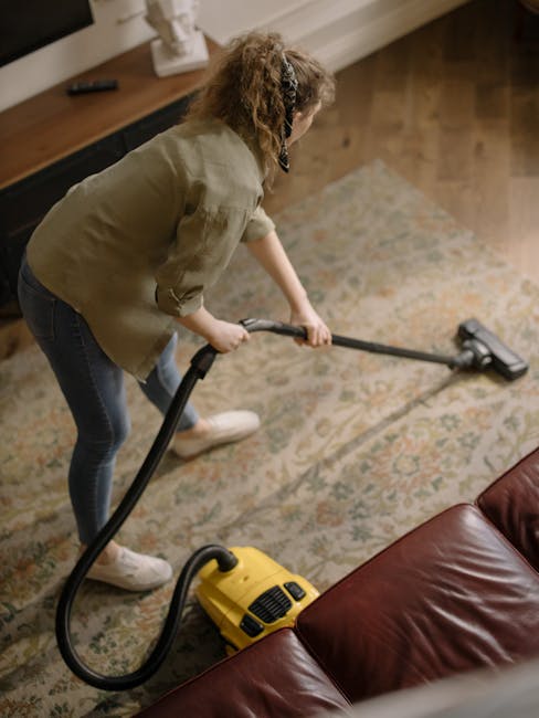 A woman engaged in domestic cleaning within a living room, using a yellow vacuum cleaner with a black hose attachment to deep clean a patterned, multi-colored carpet. She is wearing casual clothing including a beige jacket, jeans, and white sneakers, with her hair tied back, standing on a wooden floor adjacent to the carpet. The room features a leather sofa in the foreground, a wooden sideboard behind her holding a few decorative items, and is illuminated by natural light, highlighting the cleanliness and freshly maintained appearance of the carpet and surrounding surfaces. Carpet Cleaners Fulham specialises in surface cleaning and hygienic maintenance in residential spaces, focusing on thorough deep cleaning and sanitisation to ensure a hygienic environment, as seen in this scene.
