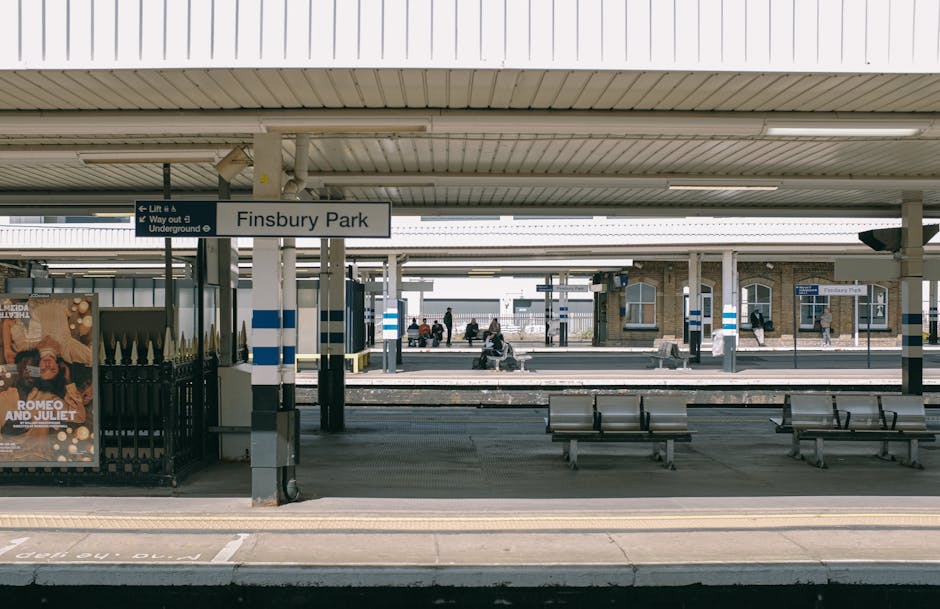 The busy interior of Fulham Broadway station, featuring a high glass and metal vaulted ceiling with intricate structural framework, large arched windows allowing natural light to illuminate the spacious concourse. The polished tiled floor appears clean and reflective, with groups of travelers walking, standing, and waiting, some pulling rolling suitcases. On the right, there are commercial kiosks and signage, including a Lush shop, with displays and advertisements visible behind glass partitions. The overall scene conveys a well-maintained, hygienic environment suited for surface cleaning, deep cleaning, and sanitisation services provided by Carpet Cleaners Fulham, capturing the station's modern architectural cleanliness and bustling atmosphere.