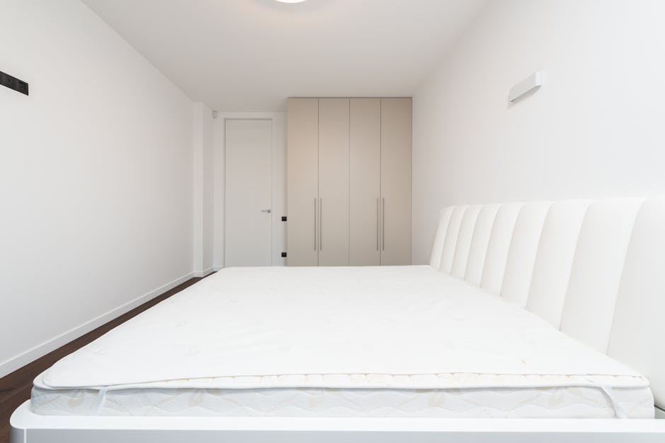 A modern bedroom featuring a grey upholstered bed with a clean, white mattress, flanked by two small grey bedside tables. The wall behind the bed has a contemporary design with vertical grey paneling 
