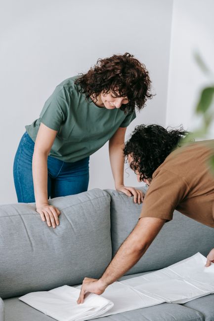 A woman with curly brown hair, dressed in a light pink blouse and white trousers, is engaged in surface cleaning on a beige upholstered sofa in a well-lit living room. She is using a white cloth to wi