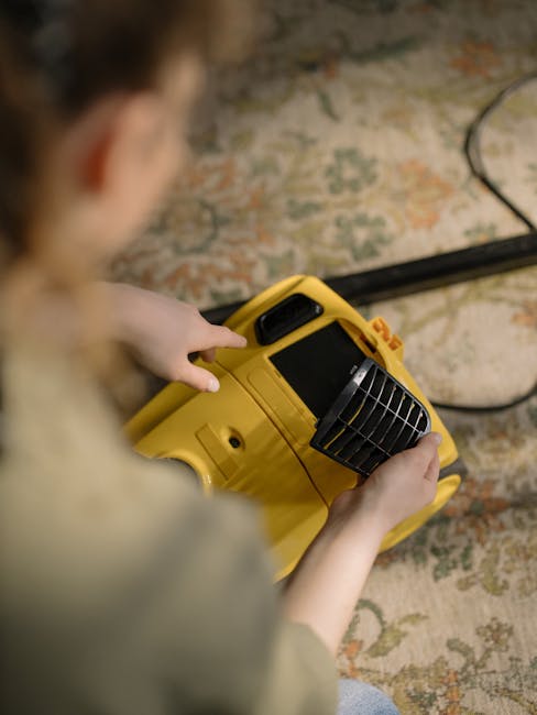 A person operating a yellow commercial-grade carpet cleaning machine with a black vent on top, positioned on a patterned area rug with floral motifs in muted pastel tones. The machine's control panel and handle are visible, and the person is pressing a button or adjusting settings. The surrounding flooring extends into a room with soft ambient lighting, and a plugged-in cord is seen trailing along the rug. The image highlights professional cleaning equipment used for surface cleaning and deep cleaning in domestic or commercial settings, with the focus on thorough hygiene maintenance. Carpet Cleaners Fulham is identified as the service provider associated with this cleaning process in Fulham, SW6, as seen from the webpage context.