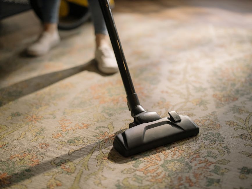 Close-up of a vacuum cleaner with a black and grey floor head actively cleaning a patterned area rug made of woven fabric, featuring floral and leafy motifs in muted tones, situated on a wooden floor in a well-lit room. The vacuum hose is connected to a long, slender wand, with a person in casual footwear visible in the background, indicating a domestic setting. The scene illustrates surface cleaning and deep cleaning as part of routine home maintenance, with the rug appearing clean and free of visible dust or debris. The image emphasizes professional cleaning techniques used by Carpet Cleaners Fulham for effective sanitisation of residential carpets in Parsons Green SW6 homes.