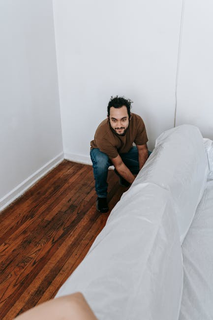 A man with curly dark hair, wearing a brown t-shirt and blue jeans, is kneeling on a polished wooden floor next to a white fabric-covered sofa in a minimalist room with plain white walls. The room is well-lit with natural light, creating a bright and clean atmosphere. The image depicts a scene related to domestic surface cleaning and maintenance, highlighting the importance of regular sanitisation and deep cleaning of furniture and flooring. The professional cleaning company, Carpet Cleaners Fulham, is associated with this scene, emphasizing quality household cleaning services on New Kings Road Fulham.