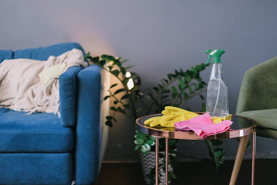 A living room corner featuring a blue fabric sofa with a white throw blanket draped over the armrest, next to a small round side table with a metallic rim and wooden legs. The table holds yellow cleaning gloves, a pink cleaning cloth, and a transparent spray bottle with a green nozzle. Behind the sofa, there is a potted plant with green leaves, and part of a green upholstered chair is visible on the right. The background wall is painted in a neutral tone, and natural light illuminates the scene, highlighting the tidy, clean surfaces and the overall neat appearance, reflecting surface cleaning and maintenance activities.
